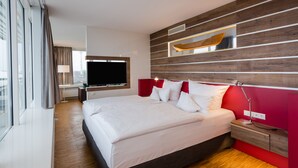 Una habitación de hotel moderna con una cama grande, un televisor de pantalla plana, un panel decorativo de madera y vistas amplias a través de grandes ventanales.