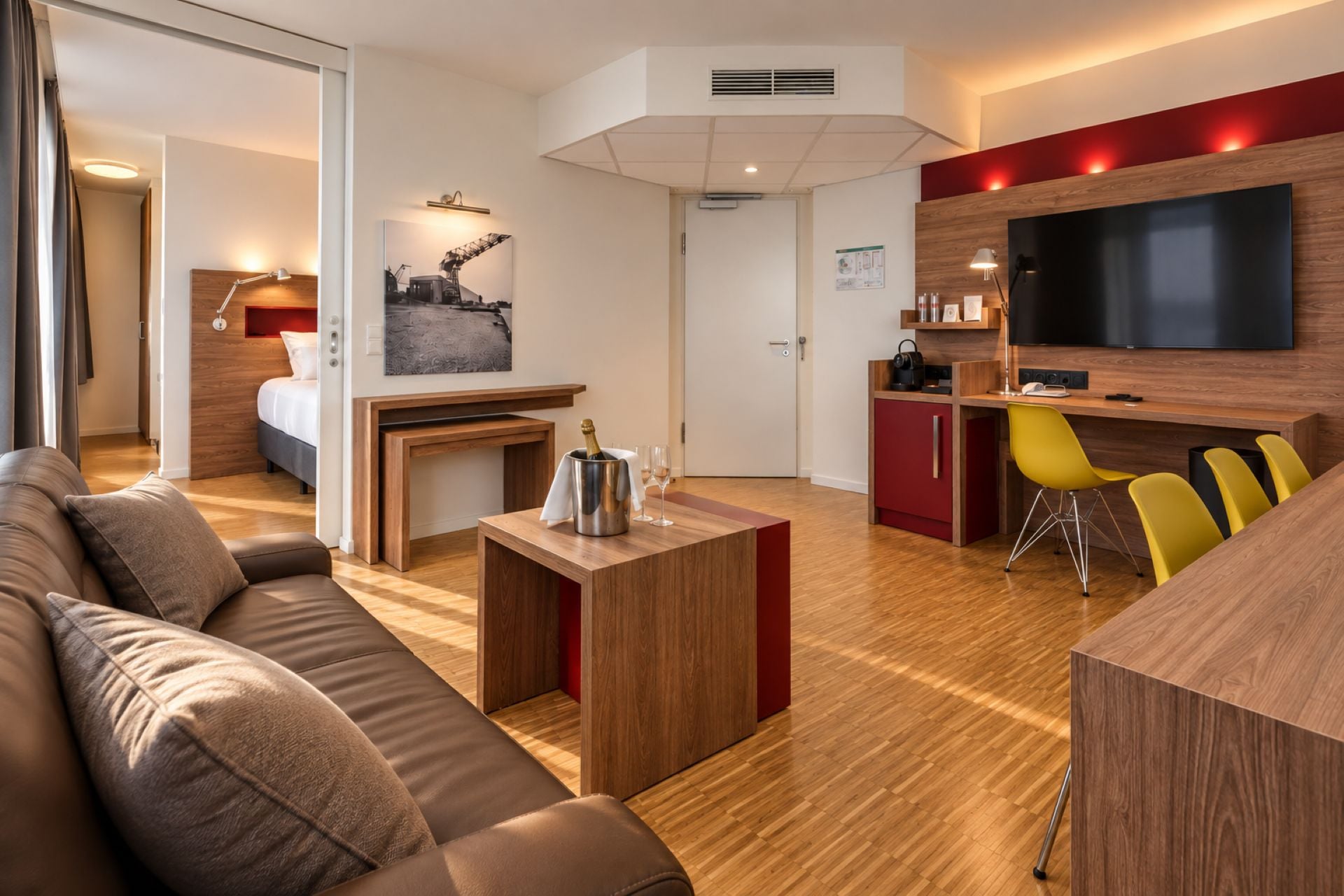 A modern hotel room with a wooden floor, a sofa, a small table with a bottle and glasses, a TV, and a red accent wall.