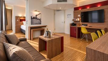 A modern hotel room with a wooden floor, a sofa, a small table with a bottle and glasses, a TV, and a red accent wall.