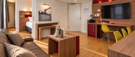 A modern hotel room with a wooden floor, a sofa, a small table with a bottle and glasses, a TV, and a red accent wall.