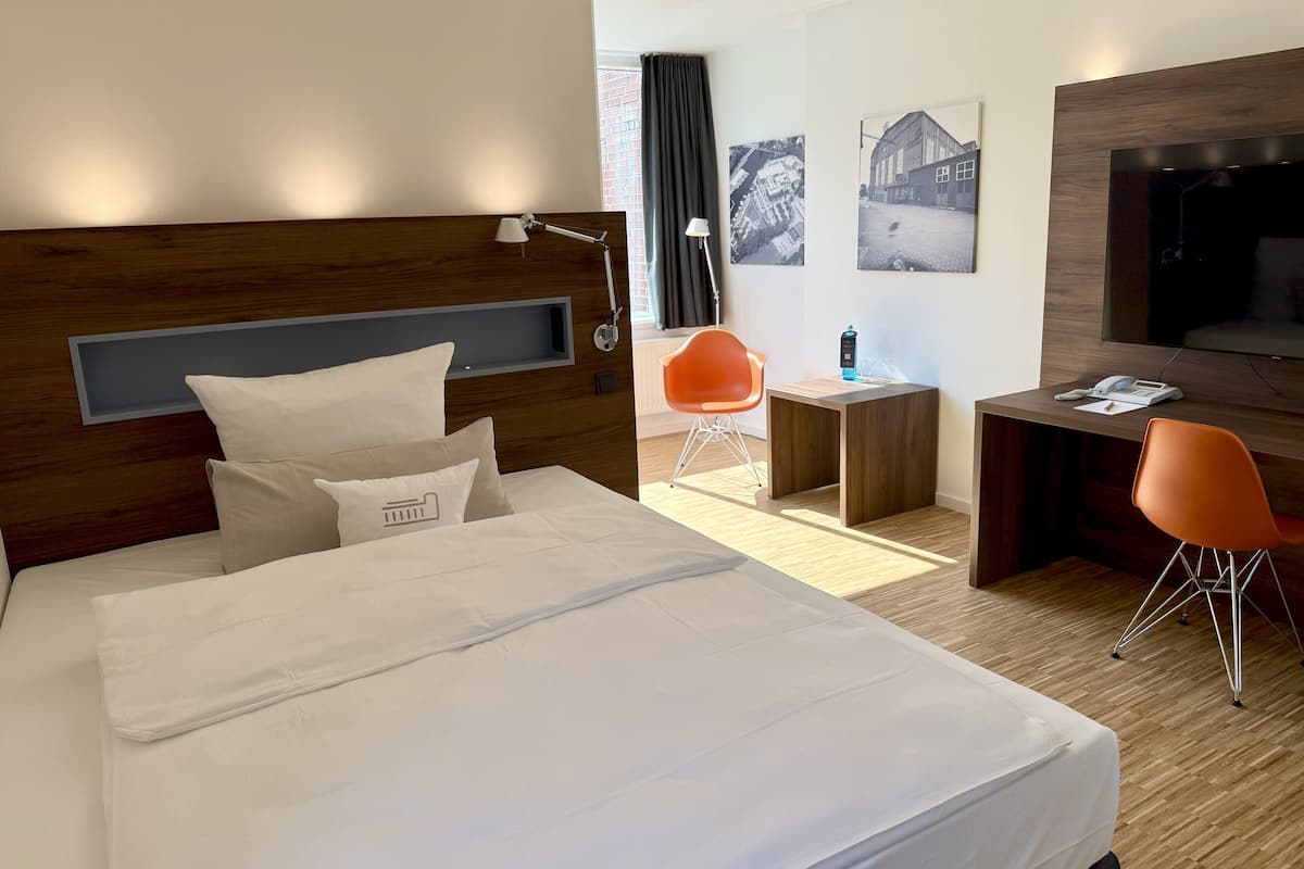 A hotel room with a large bed, a desk, a chair, a TV, and artwork on the walls.