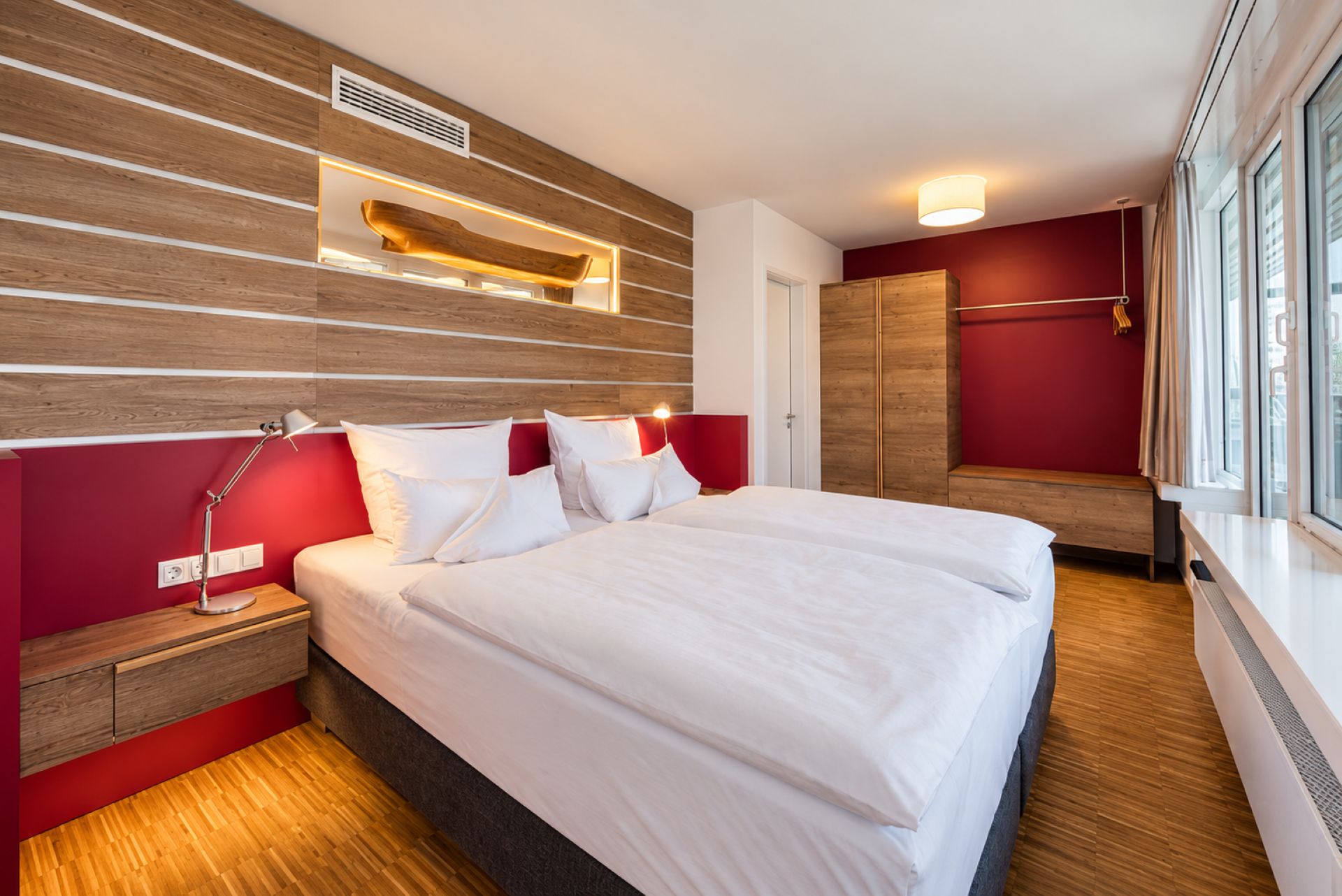 A modern hotel room with a large bed, a bedside lamp, a wooden headboard, and a window with curtains.