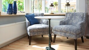 Una habitación de esquina con un sillón azul estampado, una mesita redonda y dos jarrones azules en el alféizar de una ventana.