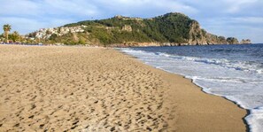 Una playa arenosa con huellas que conducen al agua, una colina rocosa con edificios al fondo y un cielo despejado.