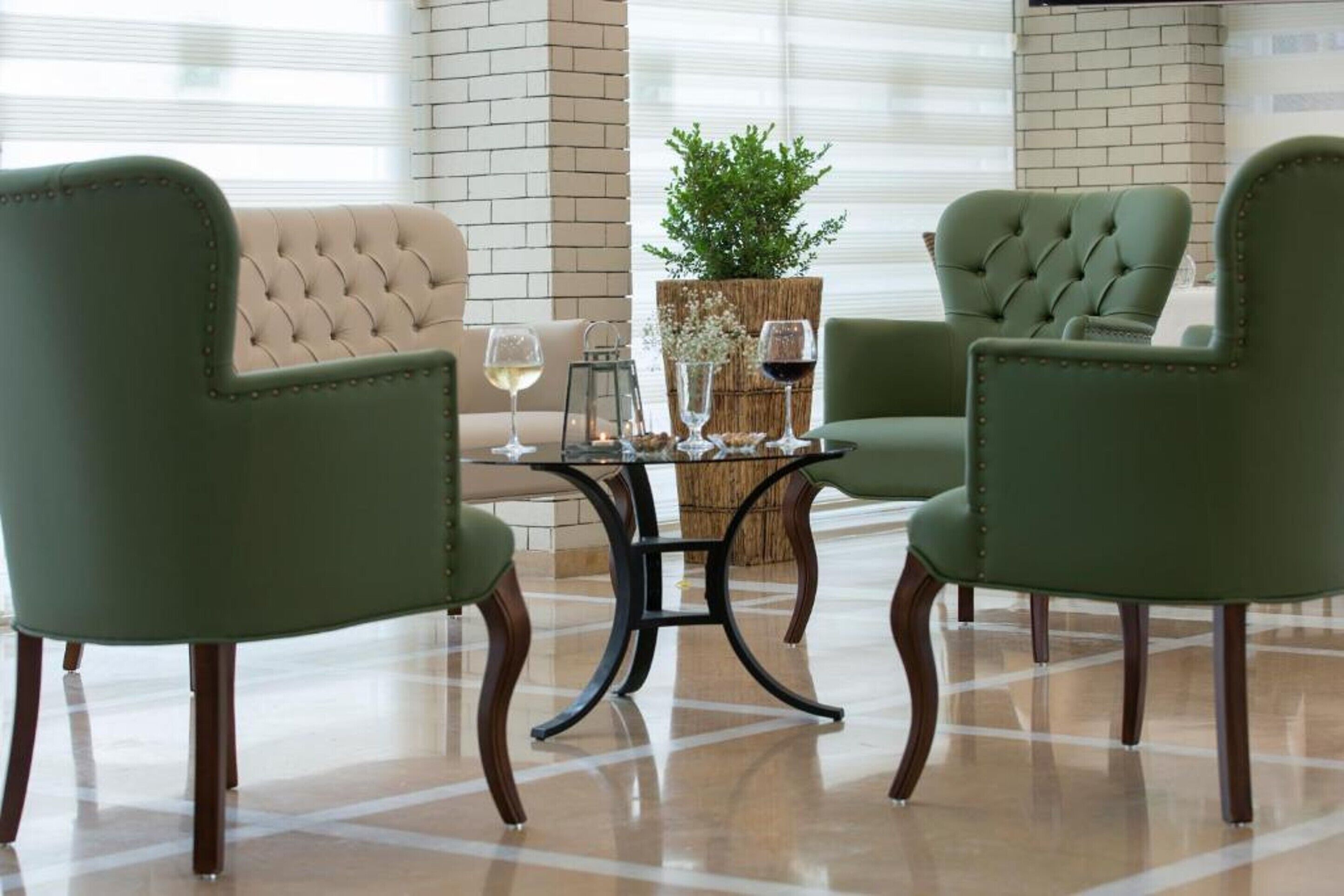 Ein Zimmer mit zwei grünen Polsterstühlen, einem kleinen Tisch mit Weingläsern und einem beigen Sofa.