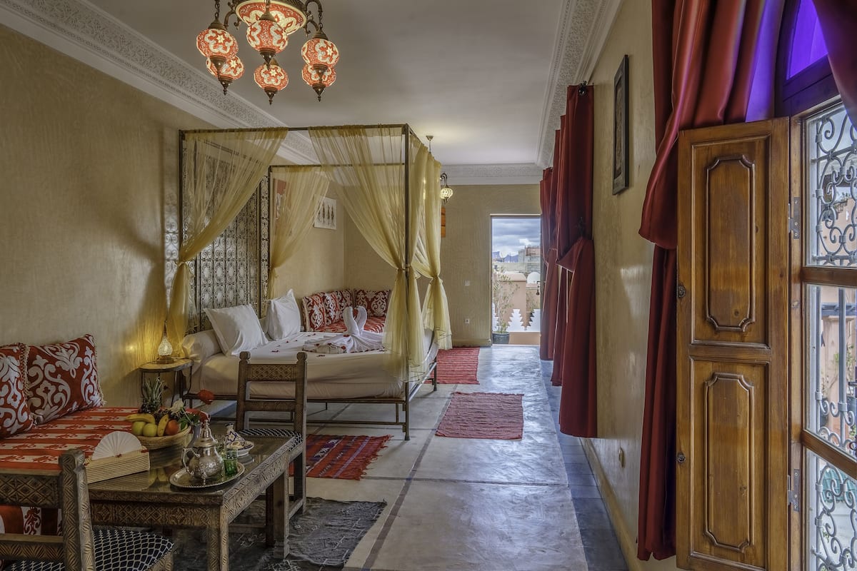 A traditional room with a four-poster bed, a dining area with a table set for two, and a view through a window with red curtains.