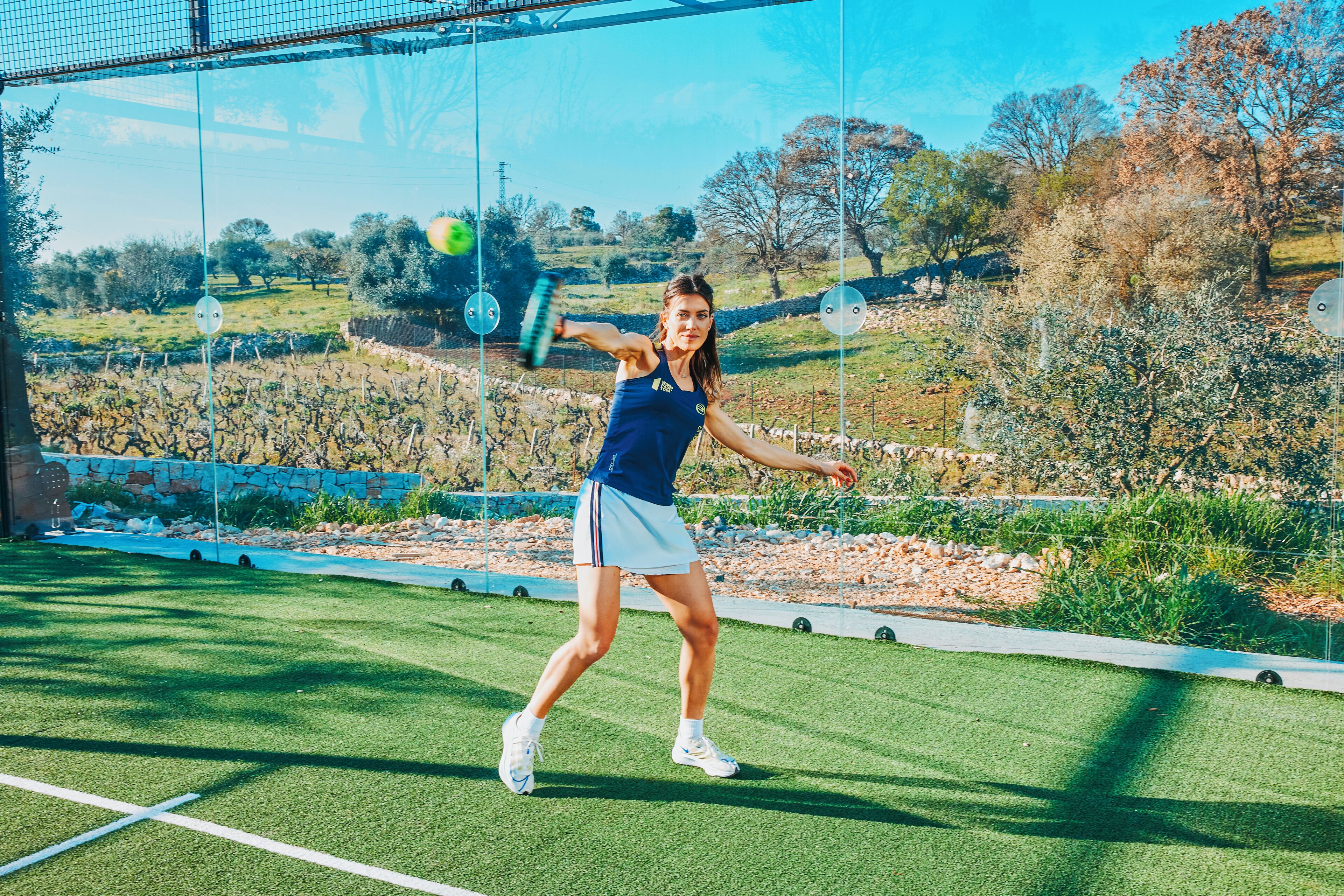 Una cancha de tenis con red, un jugador en pleno movimiento y un paisaje natural de fondo.