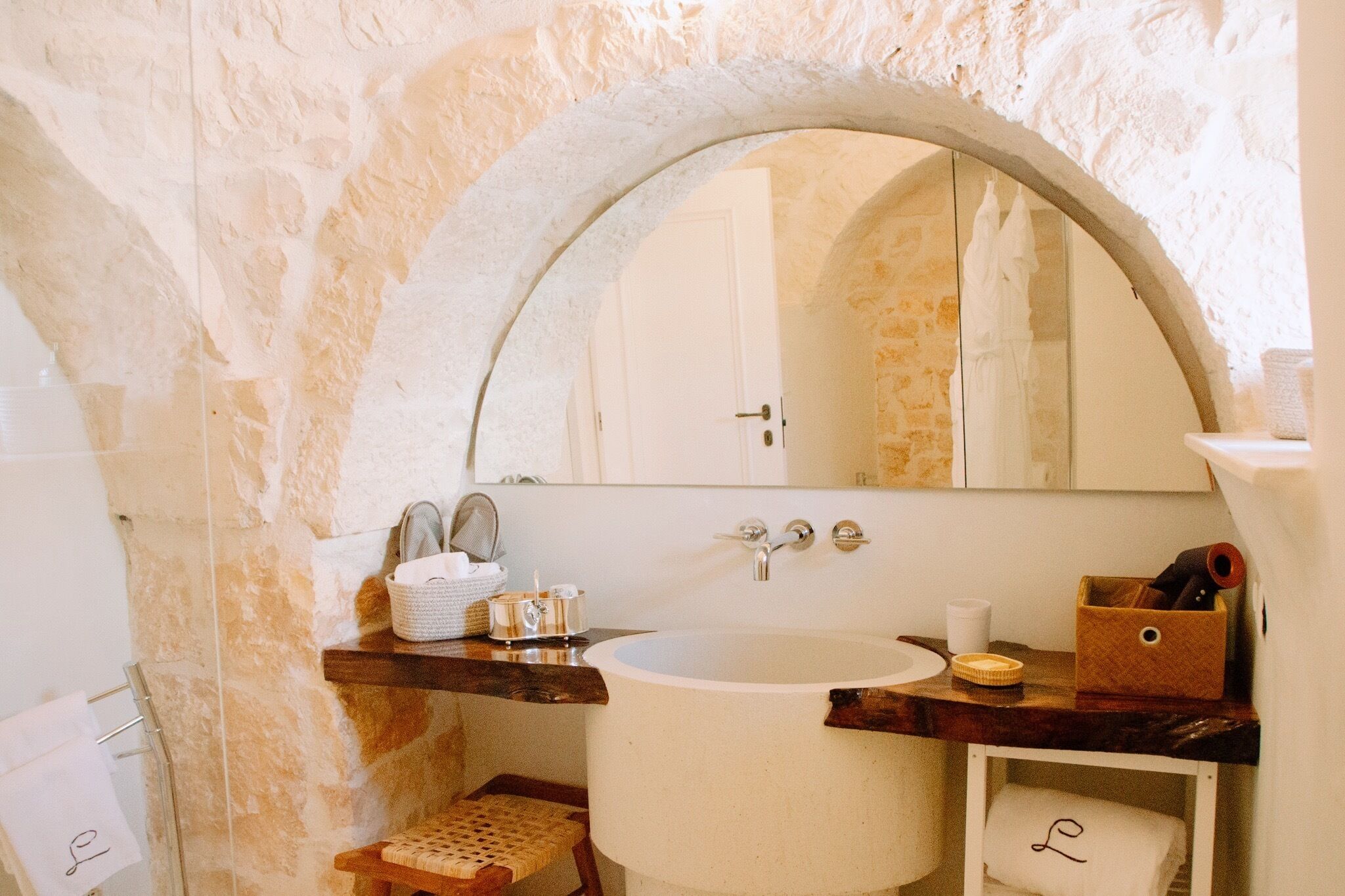 A bathroom with a stone wall, a white sink, a mirror, and a wooden shelf with toiletries.