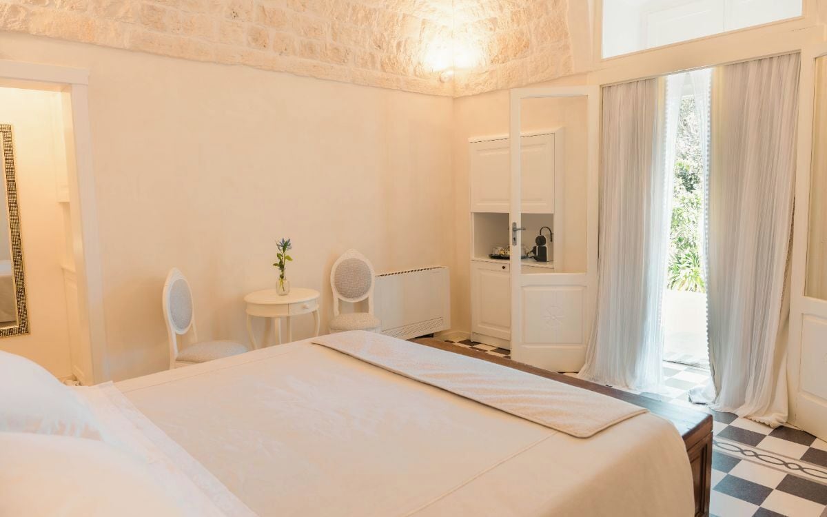 A hotel room with a large bed, a small round table with a chair, a radiator, and a view of greenery through the window.