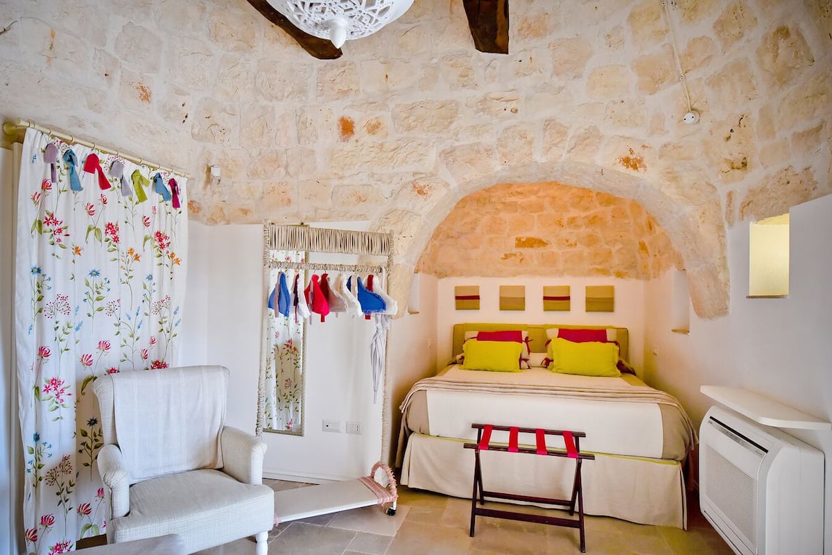 A cozy bedroom with a stone wall, a bed with yellow and red pillows, a white chair, and a small table.