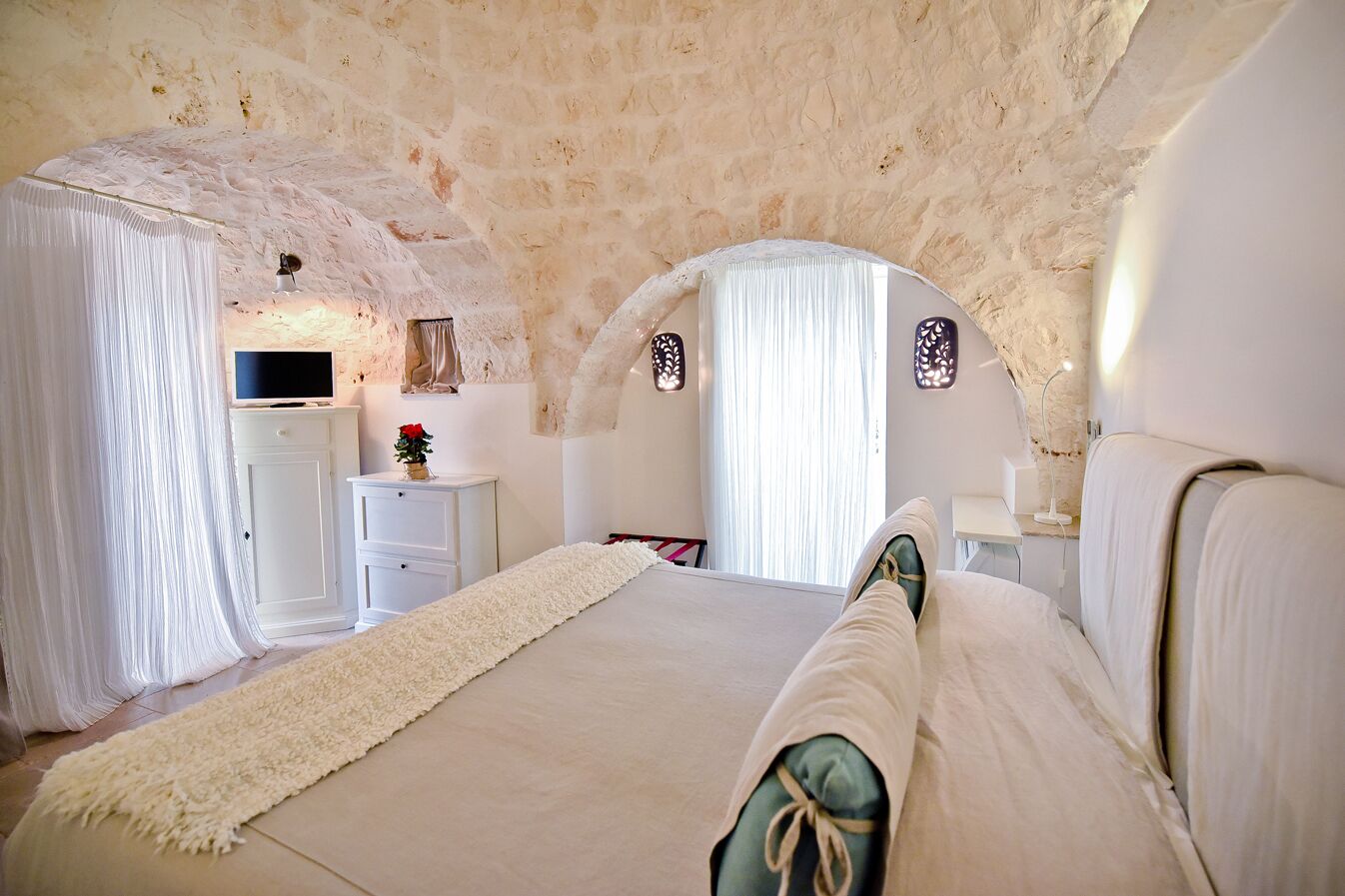 Un dormitorio con techo de piedra, una cama con ropa de cama blanca, una mesita de noche con una lámpara y una pequeña planta.