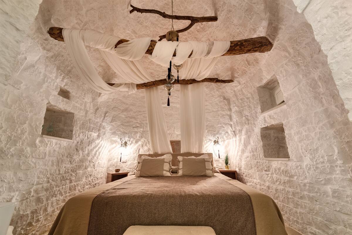 A stone-walled bedroom with a bed, draped white fabric, and a hanging light fixture.