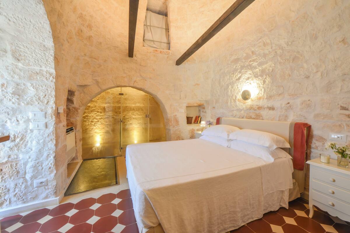 A bedroom with a stone wall, a bed, a nightstand, and a small window.
