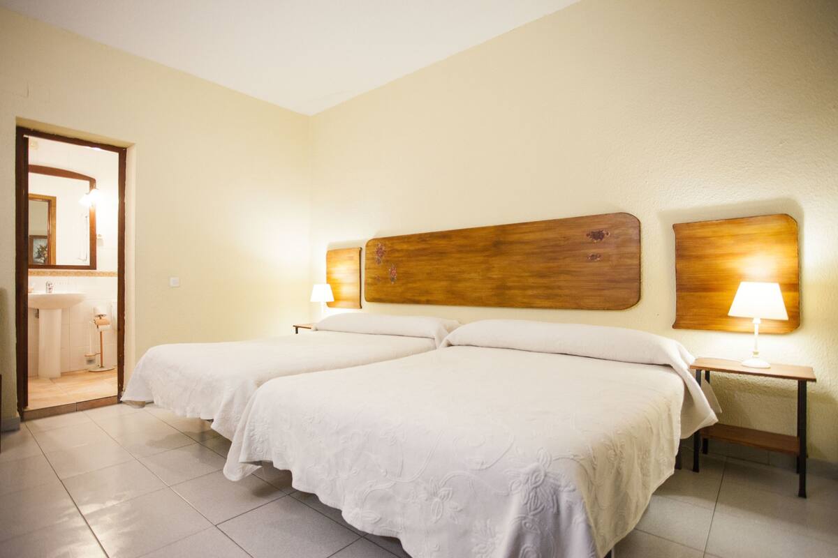 Quarto de hotel com duas camas, cabeceira de madeira, luminárias laterais e um banheiro visível por uma porta aberta.