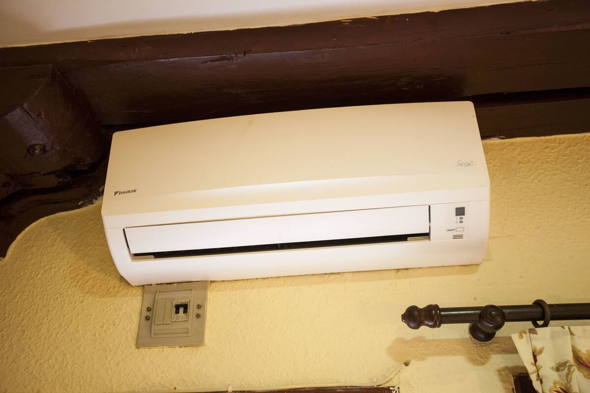 Ar condicionado Daikin branco fixado em parede texturizada, com uma tomada elétrica visível e uma barra de cortina à direita.