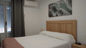 A bedroom with a wooden headboard, a bed with white bedding, a red and white striped blanket, an air conditioning unit, a painting of waves, and a window with curtains.