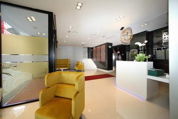 A modern hotel lobby with a reception desk, a yellow armchair, and a staircase leading to an upper floor.