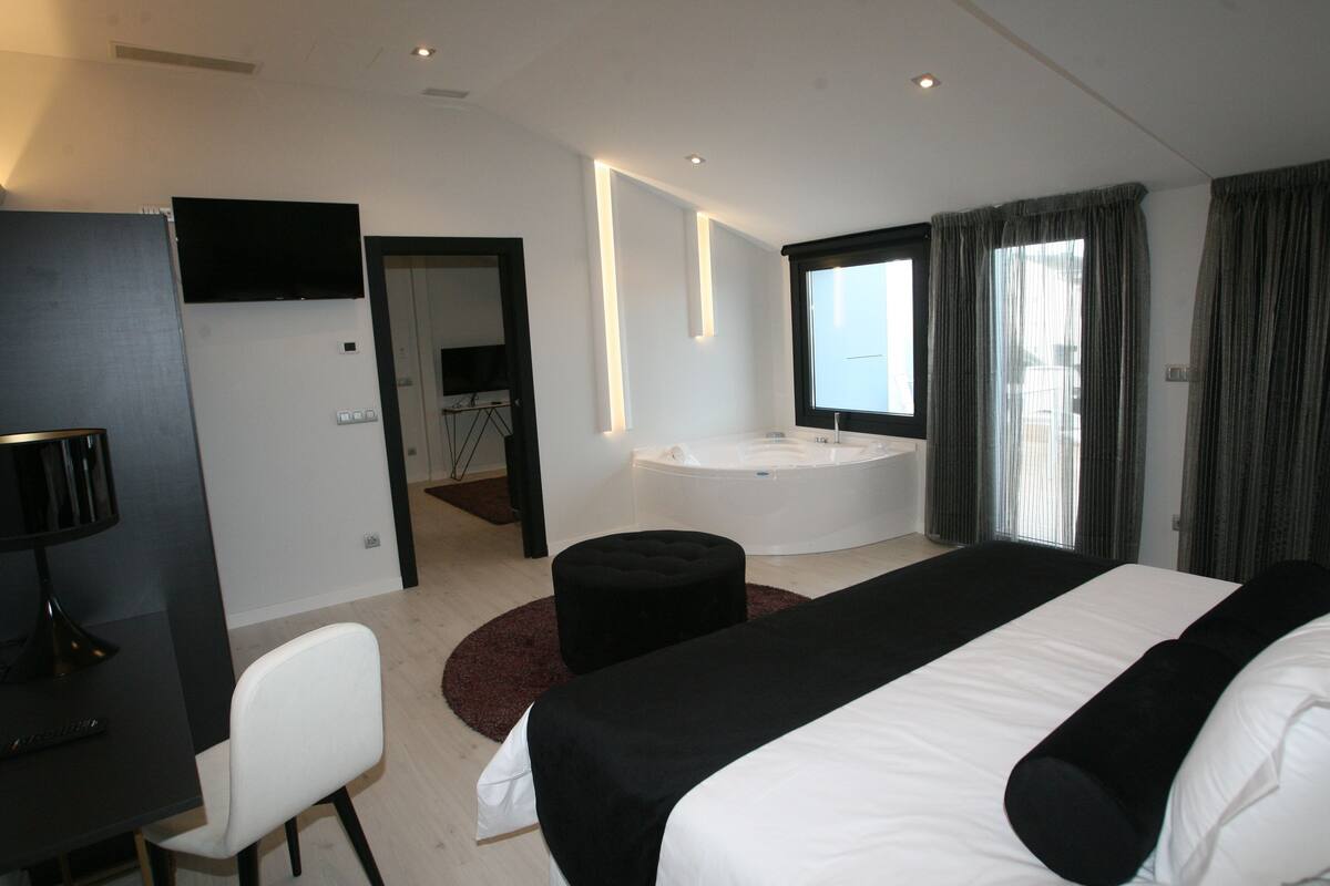 Habitación de hotel moderna con una cama grande, un escritorio, una silla, un televisor, un jacuzzi y un amplio ventanal con cortinas.
