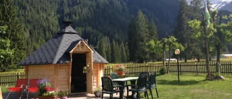 A wooden cabin with a sloped roof, a patio with a table and chairs, and a view of a forested mountain range.