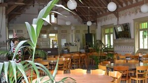 Un vaste espace dédié à la salle à manger, avec de nombreuses tables et chaises en bois, de grandes fenêtres laissant entrer la lumière naturelle et des plantes décoratives.