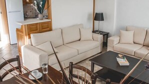 A modern living room with a beige sofa, a dining table with chairs, a coffee table, and a kitchen area in the background. - Andros Maisonettes (Andros)