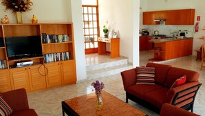 A living room with a sofa, a coffee table, and a TV. There is a kitchen area with wooden cabinets and a dining table.