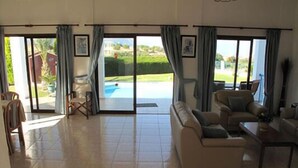 A spacious living room with a beige sofa, a dining table, and a view of a swimming pool through large glass doors.