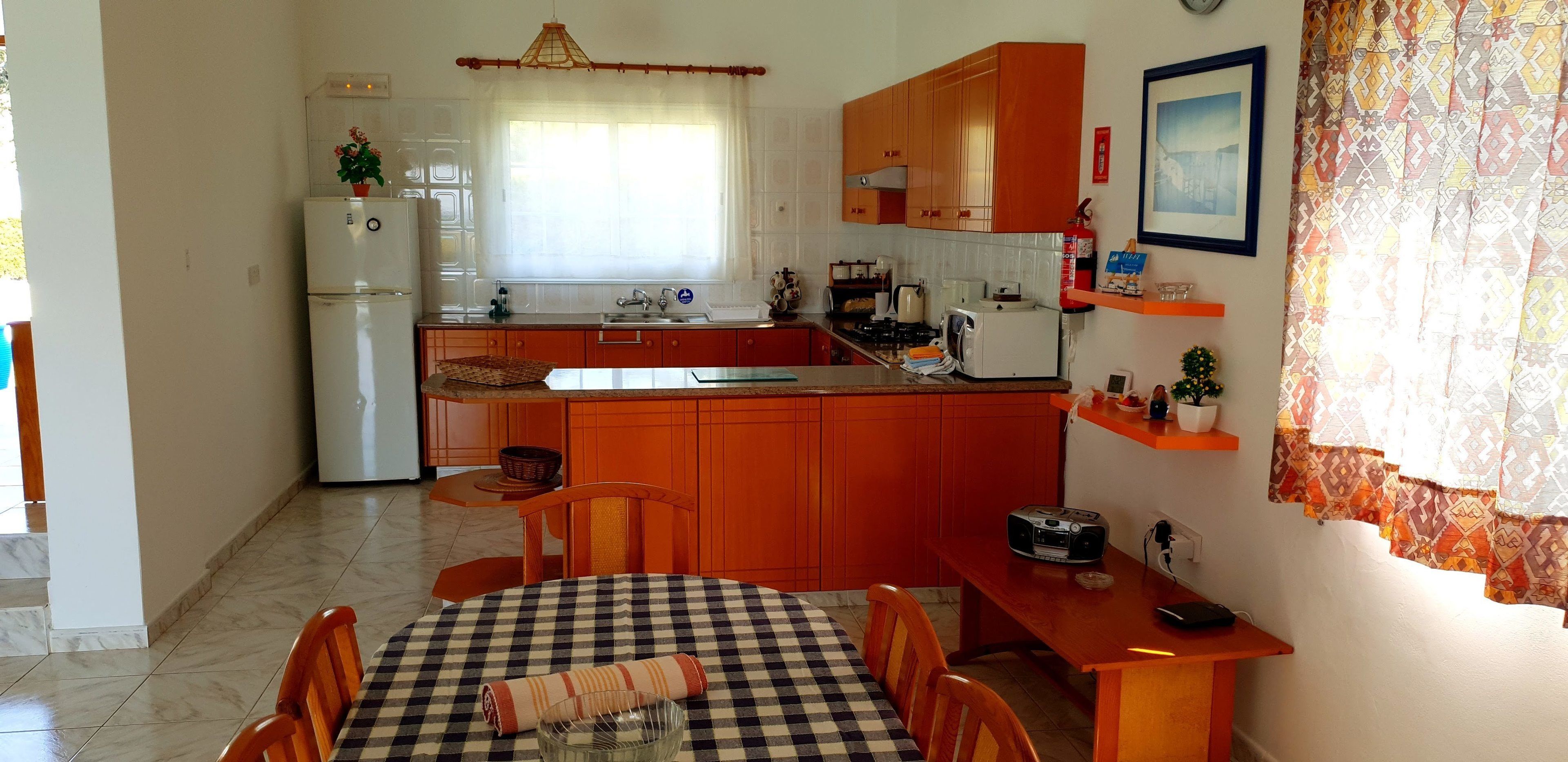 A compact kitchen with a refrigerator, sink, and stove. There is a dining table with a checkered tablecloth and chairs.