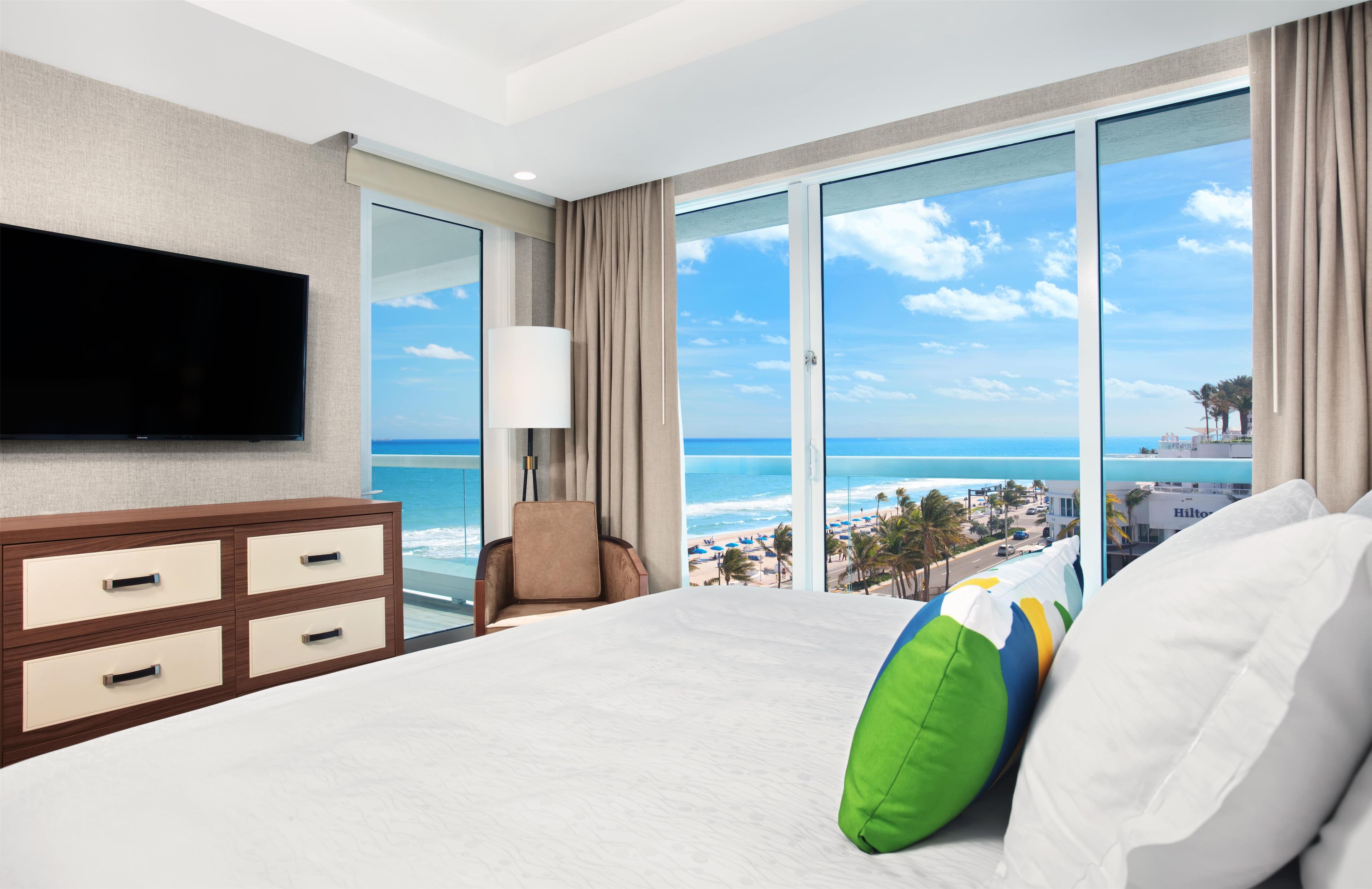 A hotel room with a large bed, a flat-screen TV, a chair, and a view of the beach.