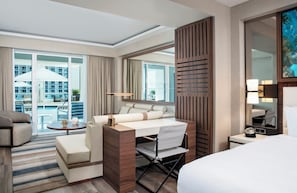 A modern hotel room with a large bed, a desk with a chair, a sofa, and a view of a pool and building through large windows. - Conrad Fort Lauderdale Beach (Fort Lauderdale)