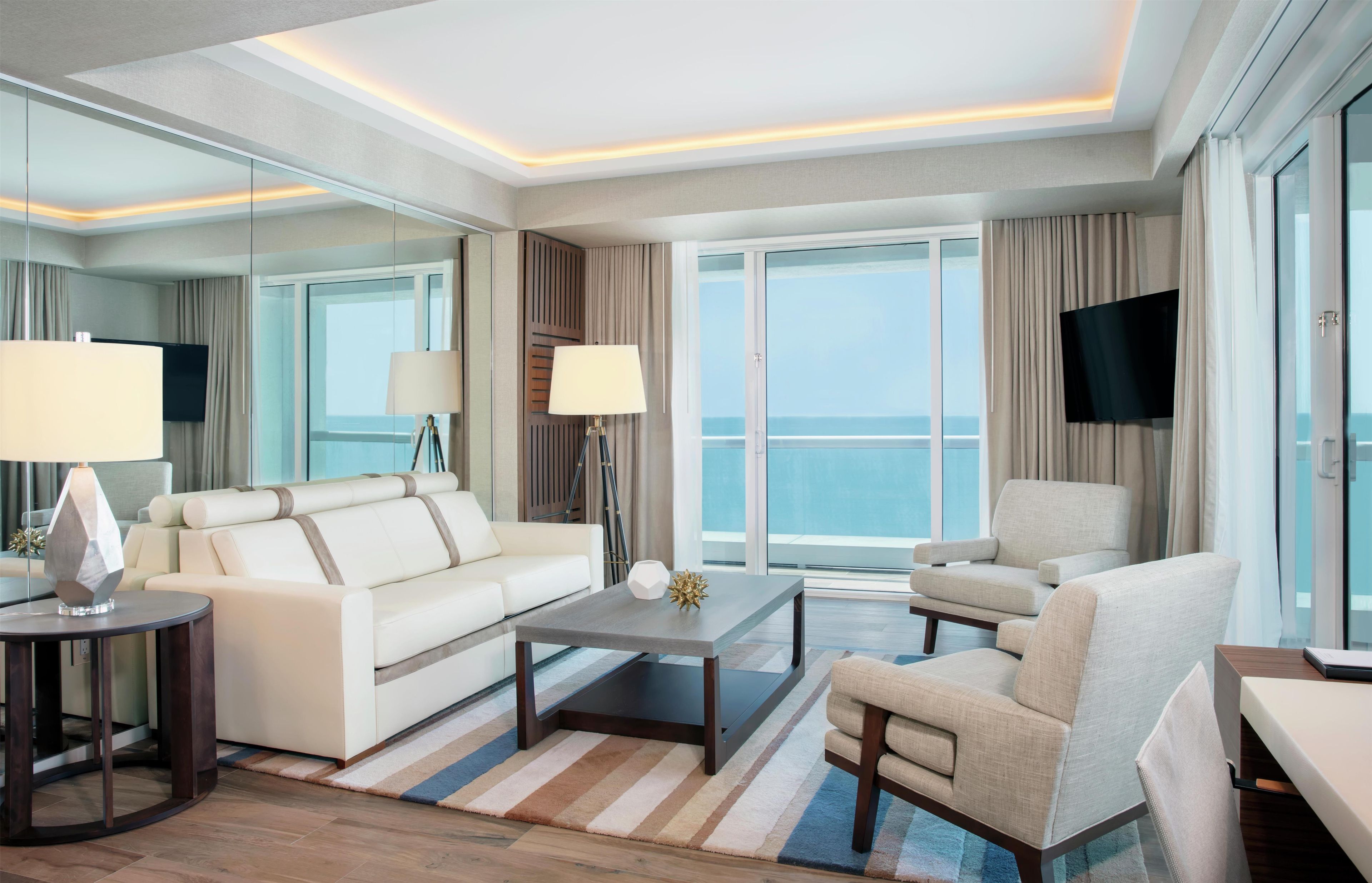 A modern hotel room with a large window, a sofa, a chair, a coffee table, and a flat-screen TV.