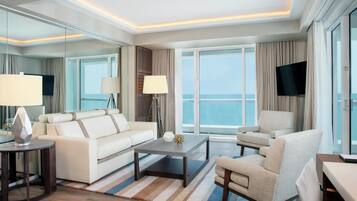 A modern hotel room with a large window, a sofa, a chair, a coffee table, and a flat-screen TV.