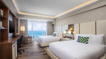 A hotel room with two beds, a desk, a TV, and a view of the sea.