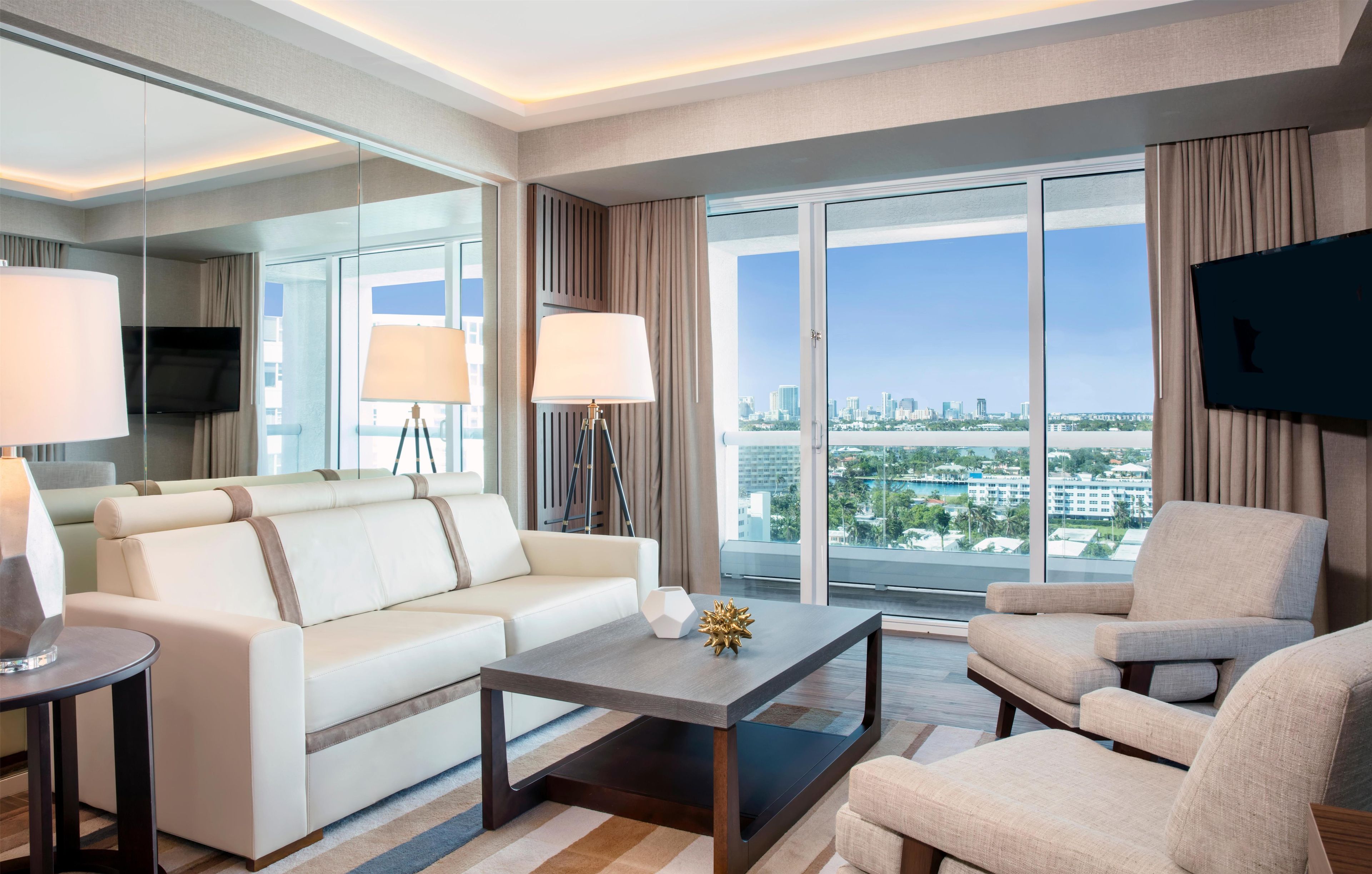 Habitación de hotel moderna con sofá, sillón, mesa de centro y vistas a la ciudad a través de amplios ventanales.