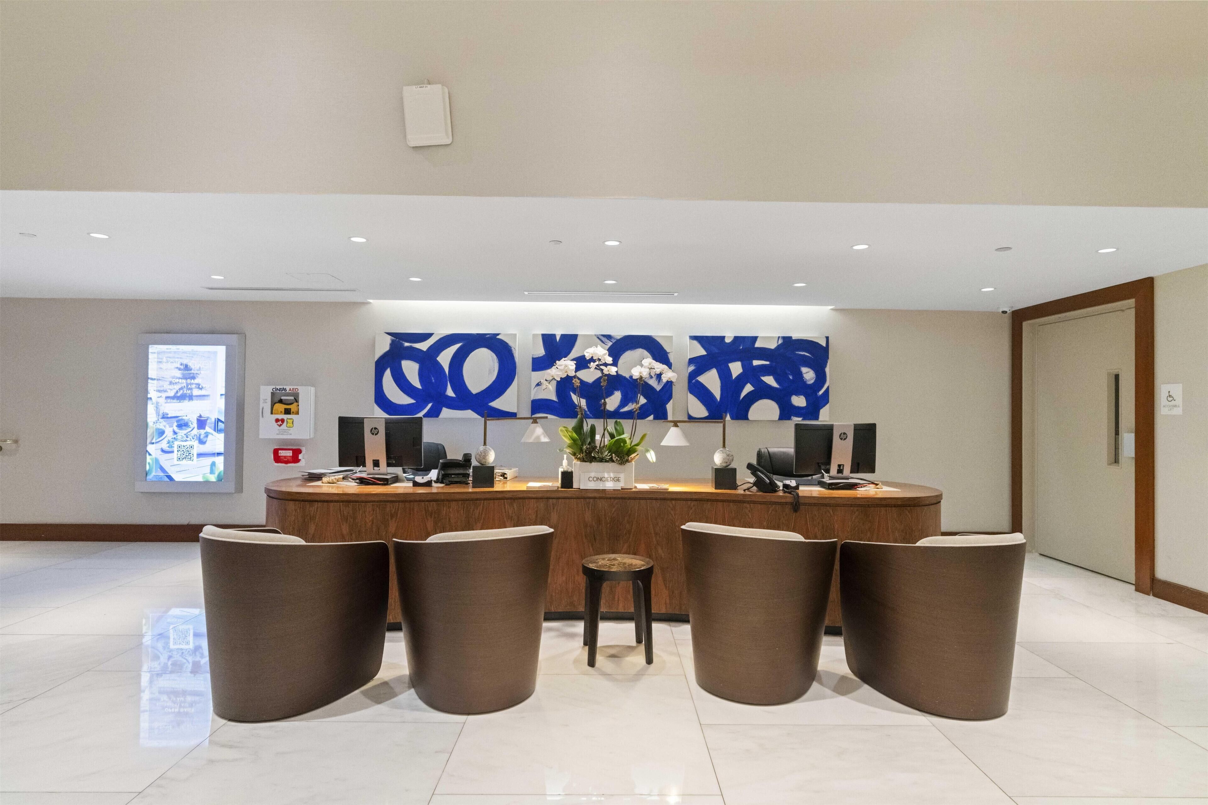 A reception area with a wooden counter, a large blue abstract artwork, a potted plant, and four brown chairs.