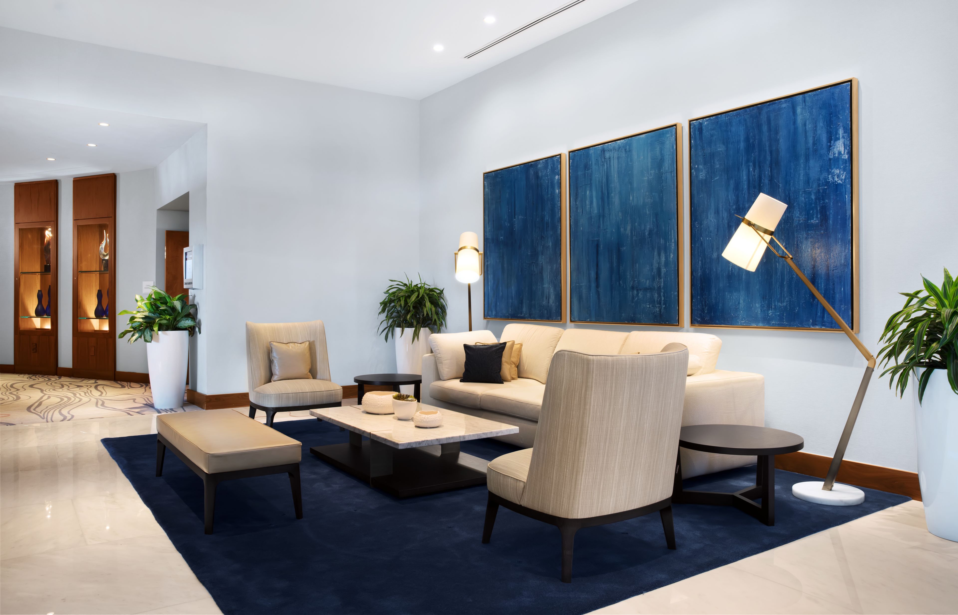 A modern lobby with a sofa, armchairs, a coffee table, and a floor lamp. There are potted plants and a large abstract painting on the wall.