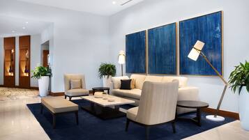 A modern lobby with a sofa, armchairs, a coffee table, and a floor lamp. There are potted plants and a large abstract painting on the wall.