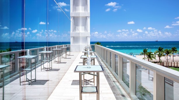 A rooftop bar with tables and chairs, overlooking a beach and ocean. - Conrad Fort Lauderdale Beach (Fort Lauderdale)