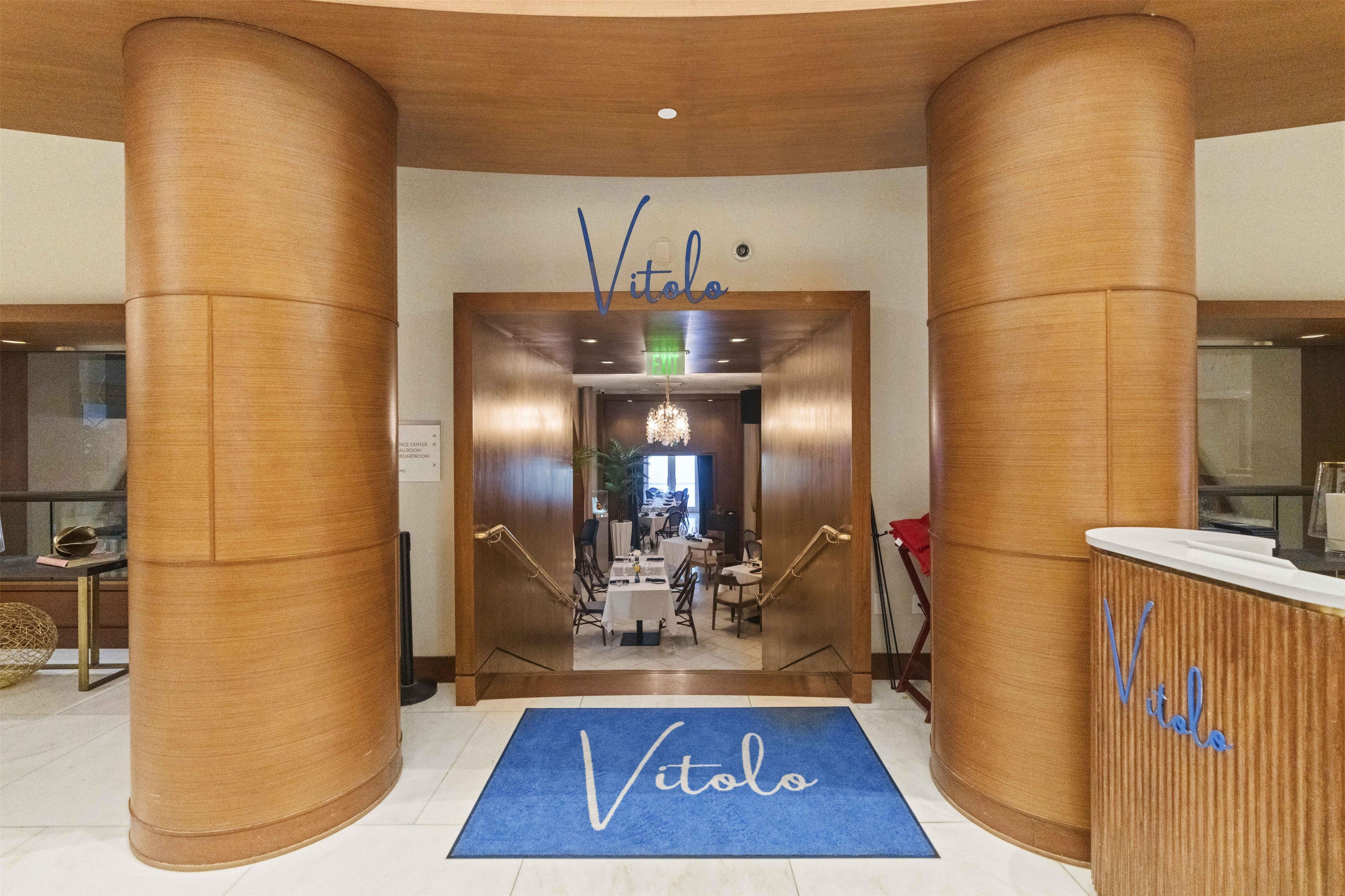A hotel entrance with a reception desk, a blue mat with the hotel's name, and a staircase leading to the dining area.