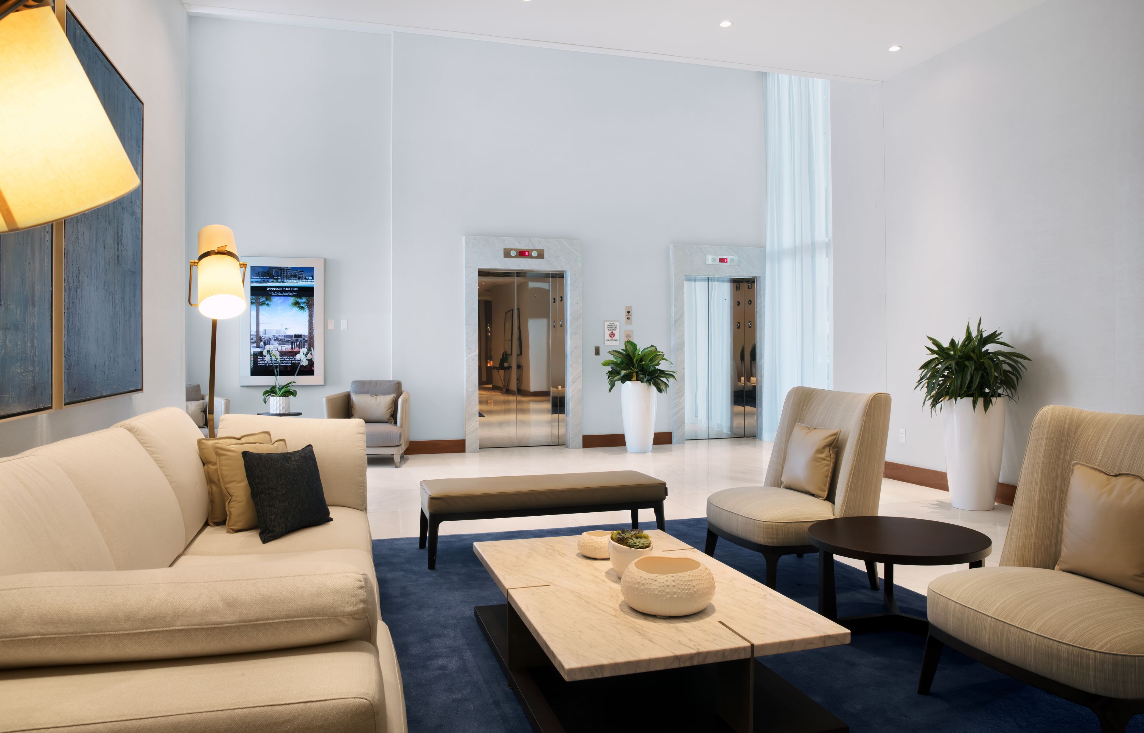 A modern lobby with a beige sofa, a coffee table, and two armchairs. There are potted plants and a large mirror on the walls.