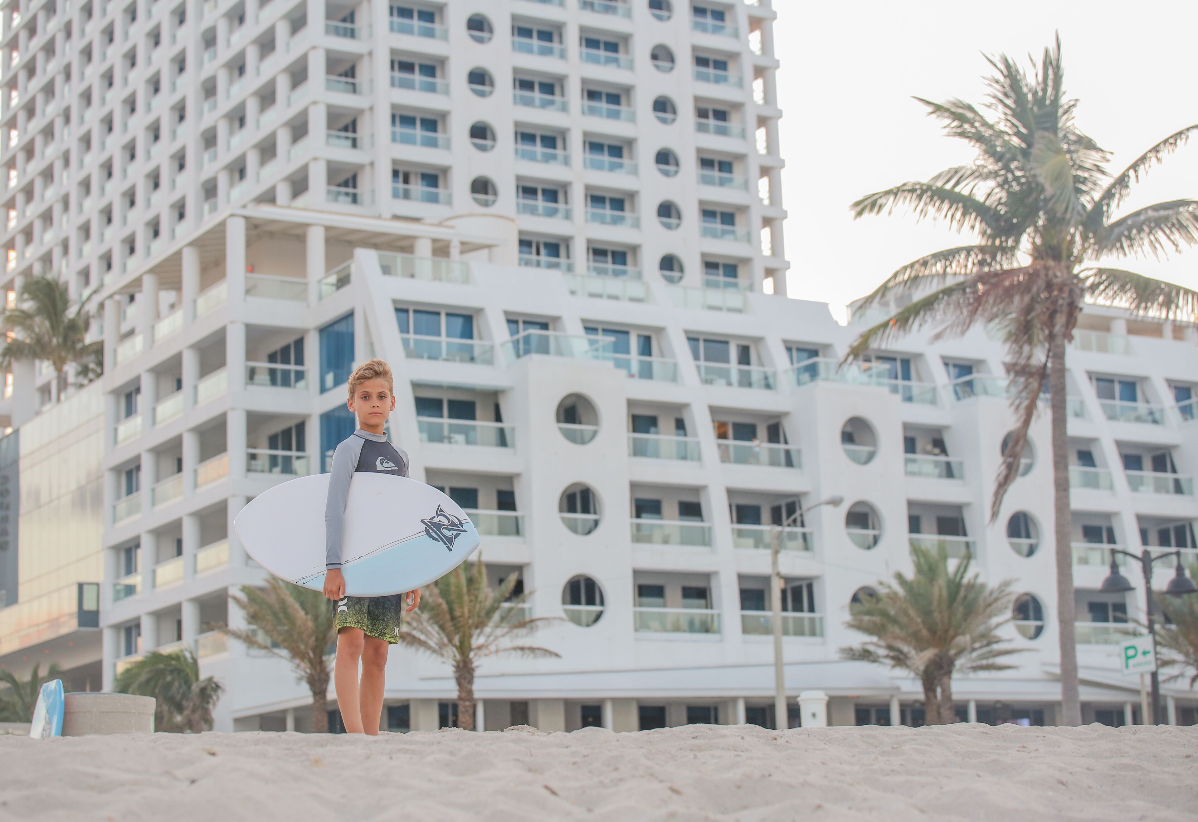 A person holding a surfboard in front of a modern building with circular windows and palm trees.