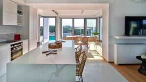 A modern kitchen with a large island, dining area with wicker chairs, and a TV on the wall. - Empire Luxury Villas (Paros)