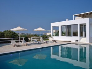 A modern pool area with lounge chairs and umbrellas, a clear blue pool, and a white building with large windows.