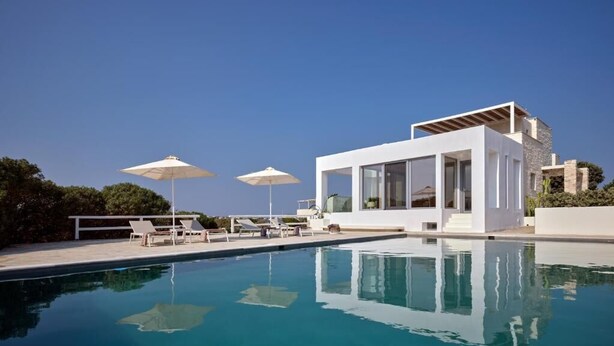 A modern poolside area with a clear blue pool, white lounge chairs, and umbrellas. - Empire Luxury Villas (Paros)