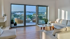 A modern living room with a large sliding glass door leading to a balcony, a white sofa, a coffee table, and a view of the city. - Empire Luxury Villas (Paros)