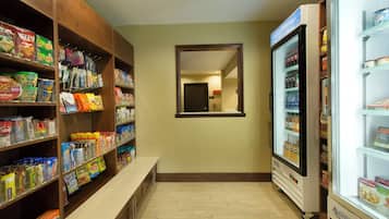 A hotel pantry with shelves stocked with snacks and beverages, a mirror on the wall, and a refrigerator.
