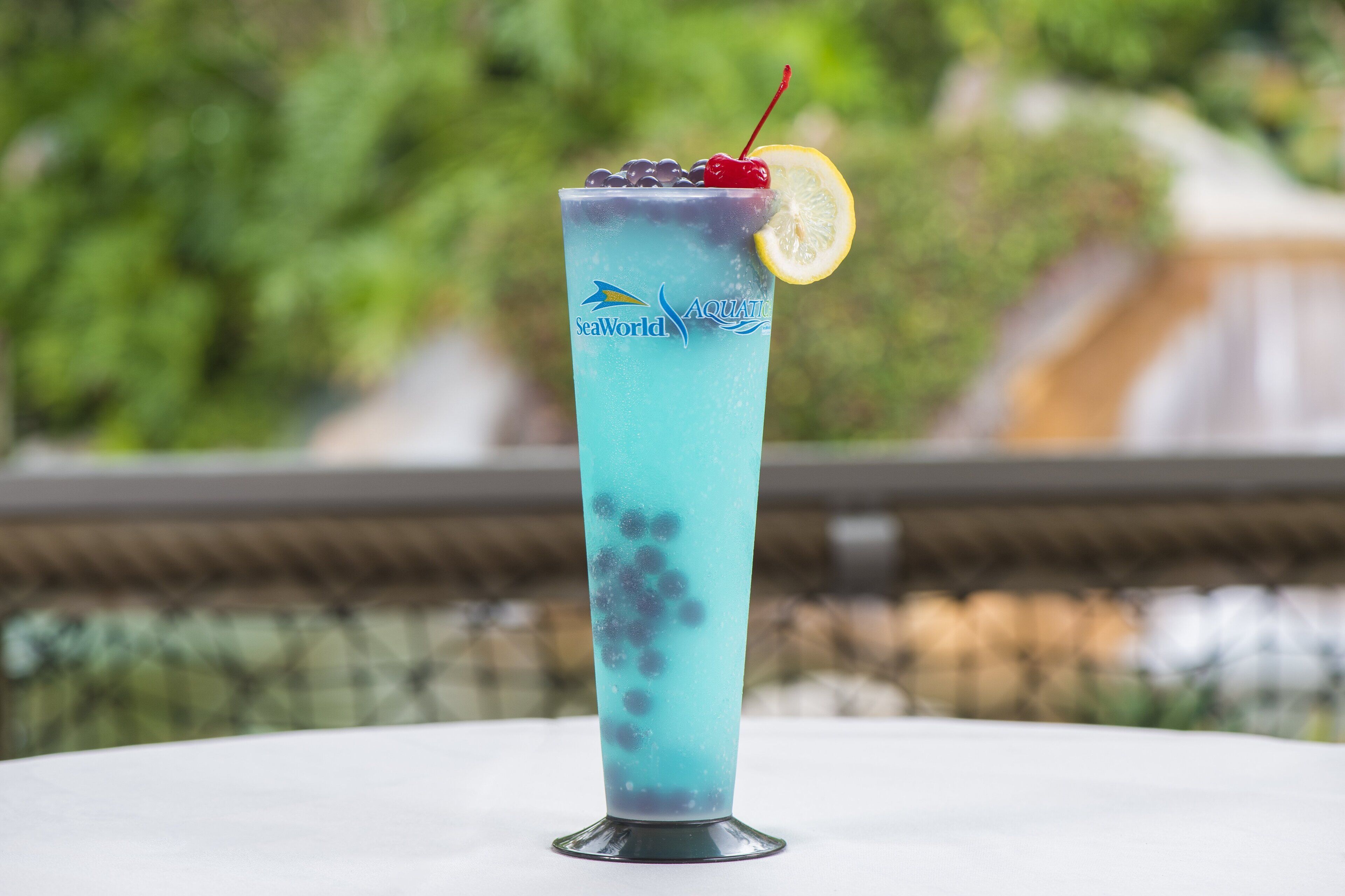 Un vaso alto con una bebida azul, una cereza y una rodaja de limón sobre una mesa.
