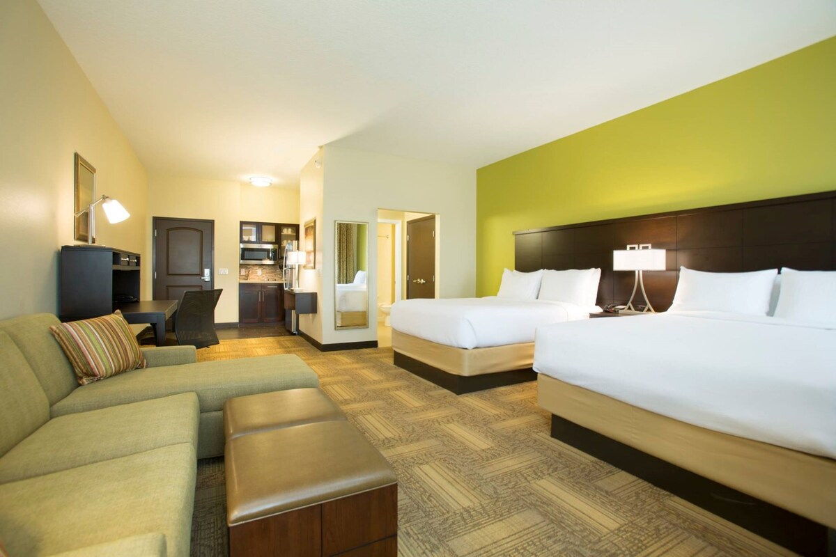 A hotel room with a large bed, a sofa, a small desk, and a kitchenette.