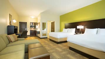 A hotel room with a large bed, a sofa, a small desk, and a kitchenette.