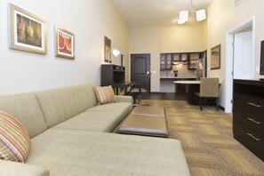 A living room with a sofa, ottoman, and a dining area with a table and chairs. There is a kitchen in the background with cabinets and a refrigerator.